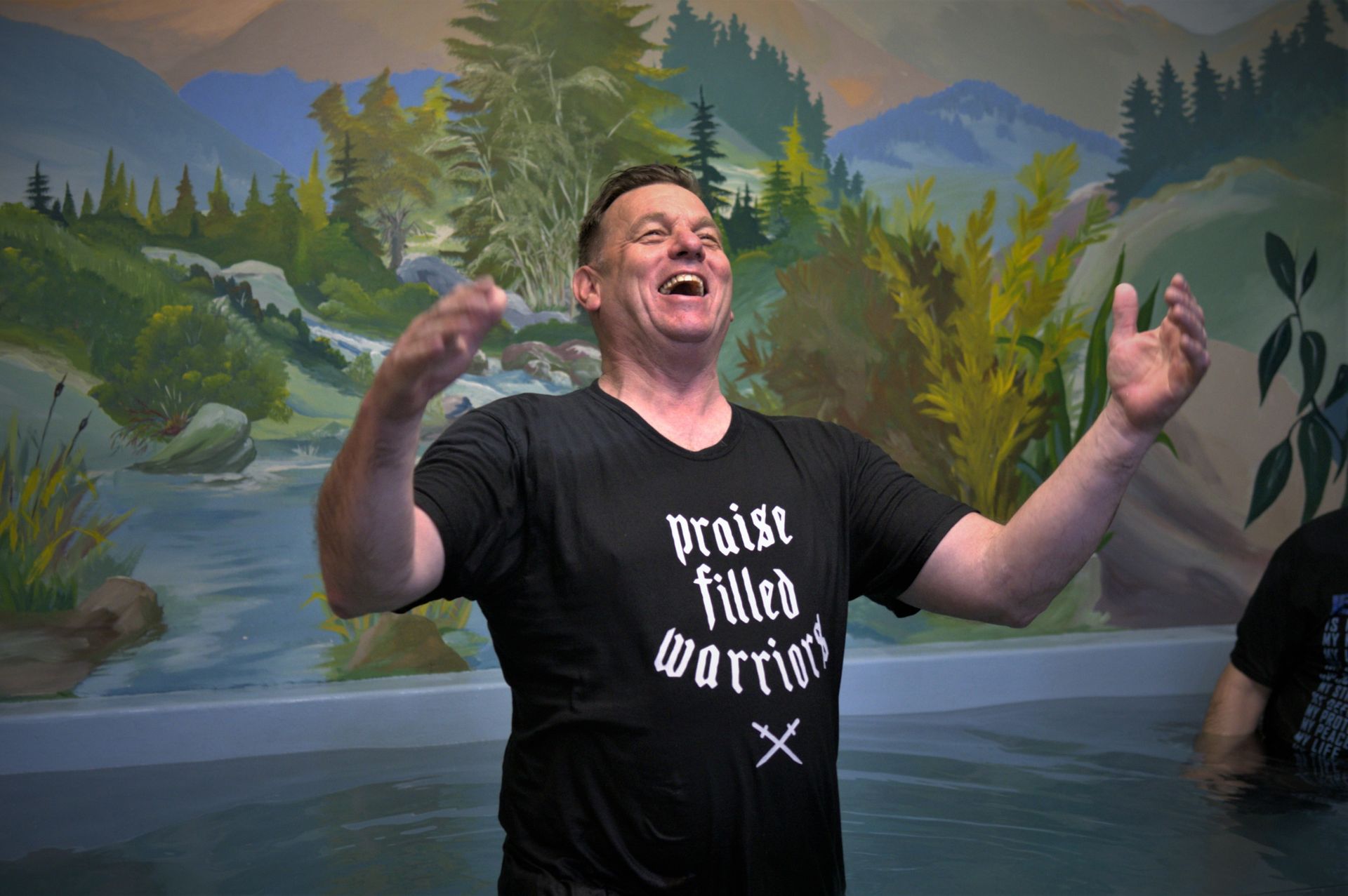 A man in a black shirt is standing in the water with his arms outstretched.