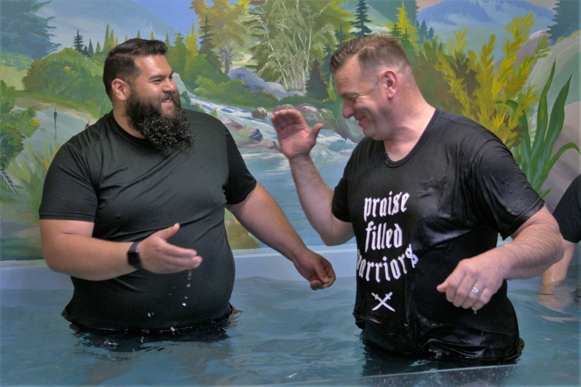 Two men are standing in the water and one of them is wearing a shirt that says `` praise filled warriors ''.