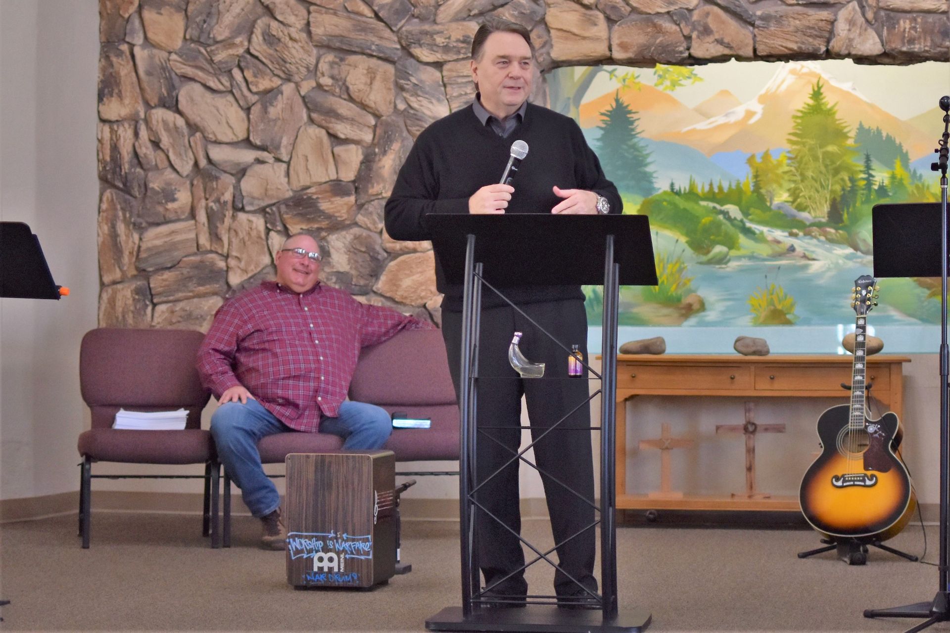 A man is standing at a podium talking into a microphone while another man sits on a couch.