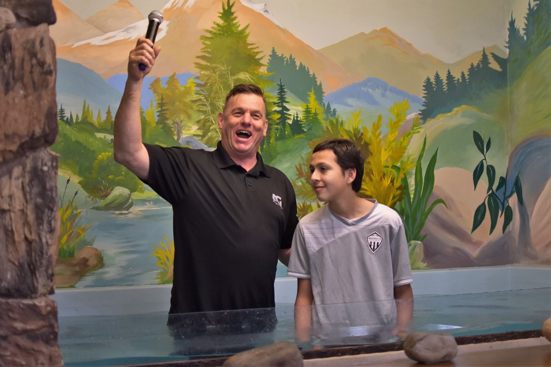 A man is holding a microphone while standing next to a boy in the water.