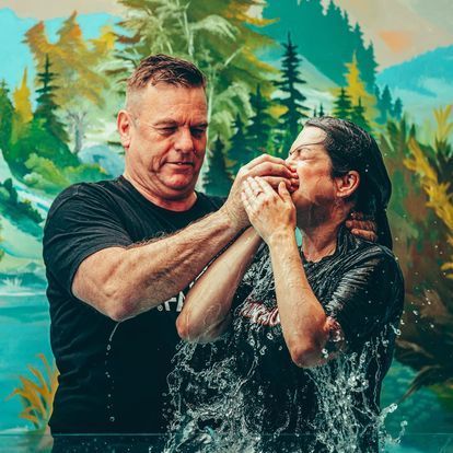 a man is baptising a woman in front of a painting of trees .