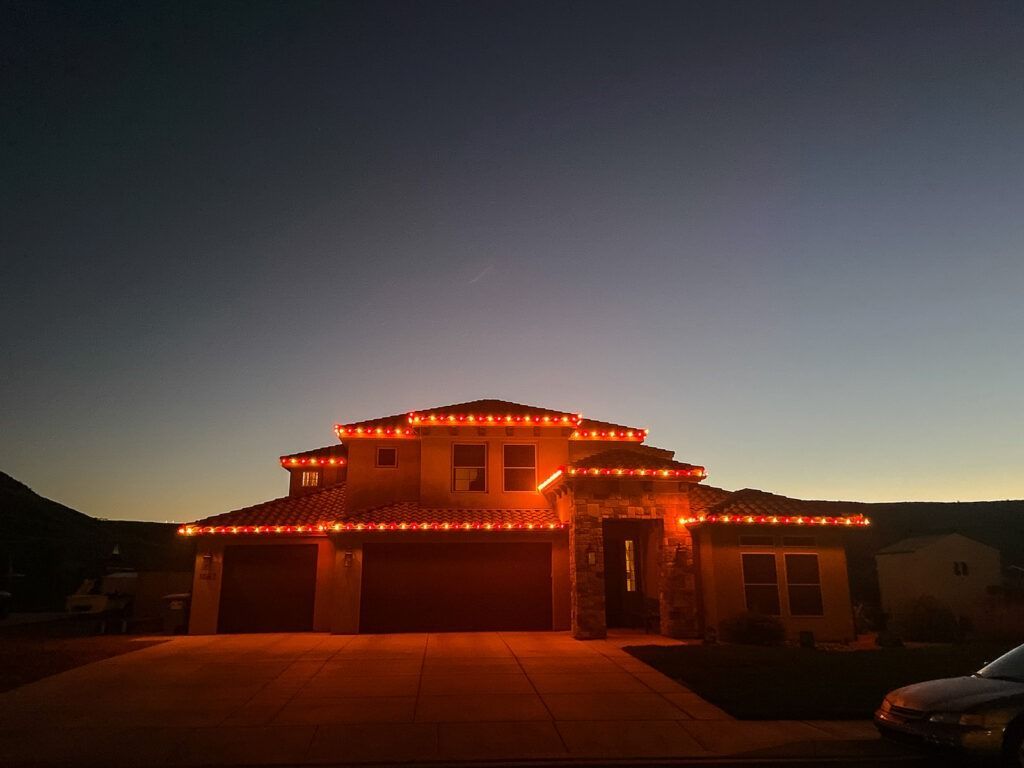 A two-story house at dusk outlined with glowing orange festive lights across its rooflines and garage.