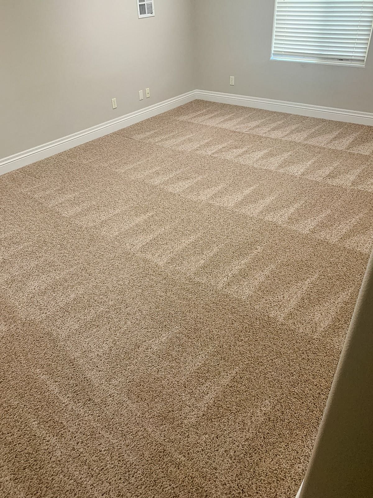 A wide shot of an empty room with tan, textured carpeting, light beige walls, white trim, and a window with blinds.
