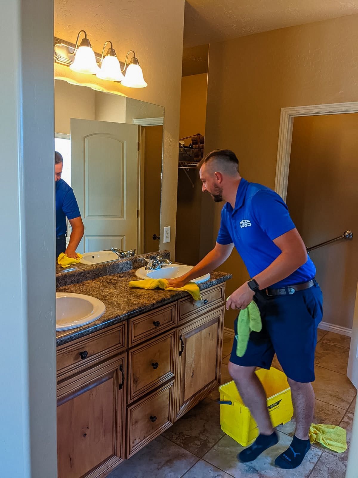A person in a blue uniform cleaning a bathroom countertop with a yellow cloth.