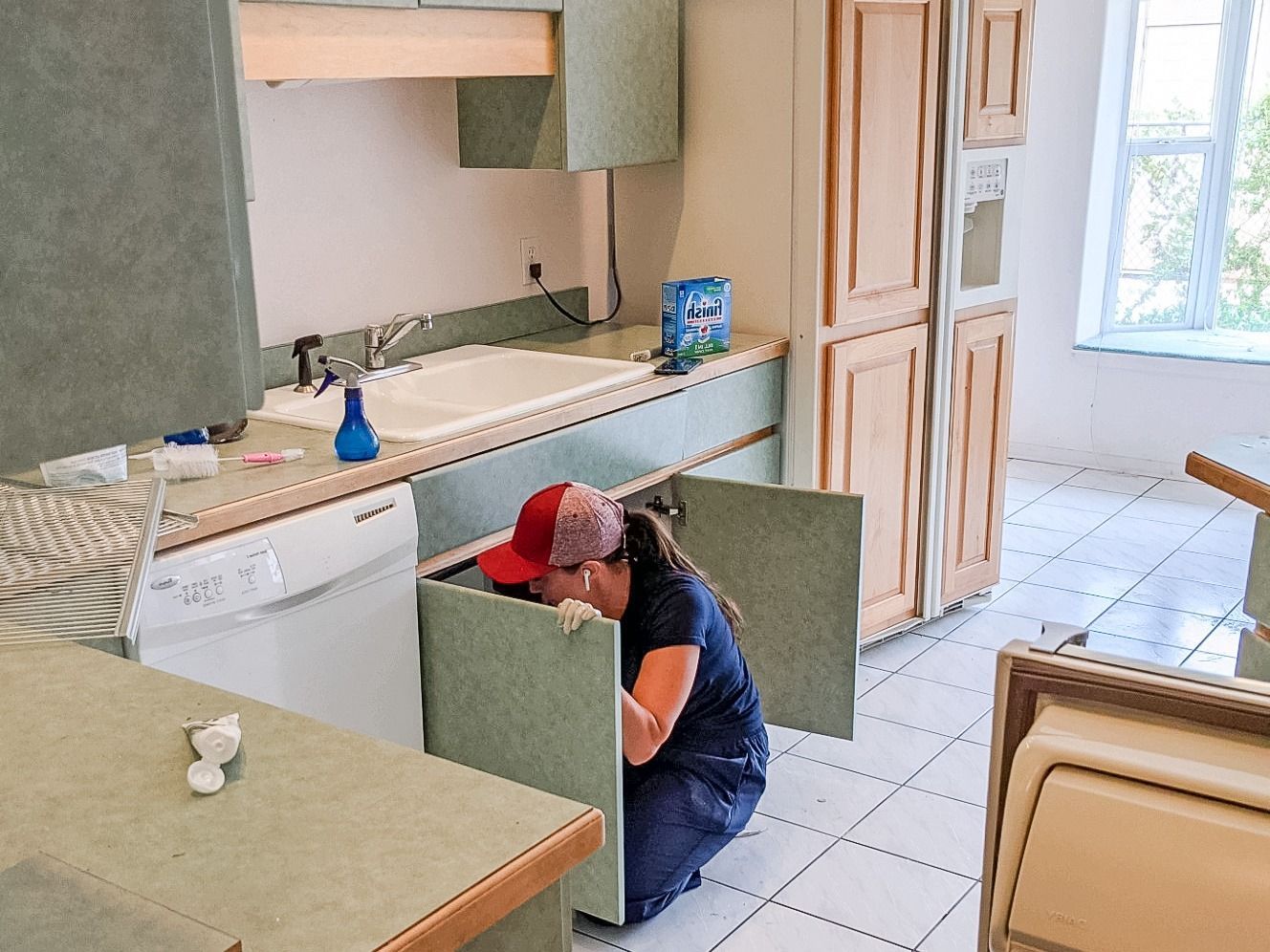 A person wearing a red baseball cap kneels on a tiled kitchen floor, working inside an open cabinet under the sink.