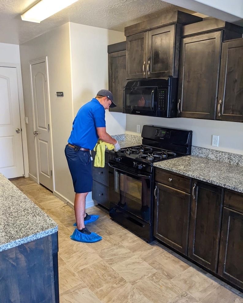 A person wearing blue shoe covers and a blue shirt cleans a black gas stovetop in a kitchen with dark wood cabinets.