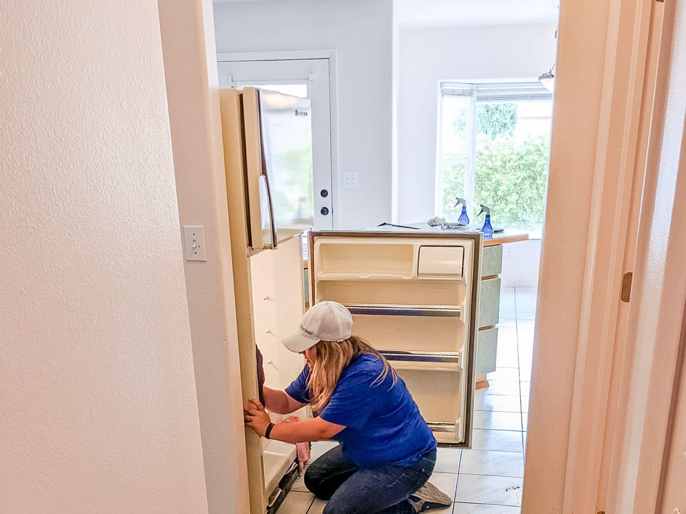 A person in a blue shirt kneels on a tiled floor while cleaning or working inside an empty refrigerator.