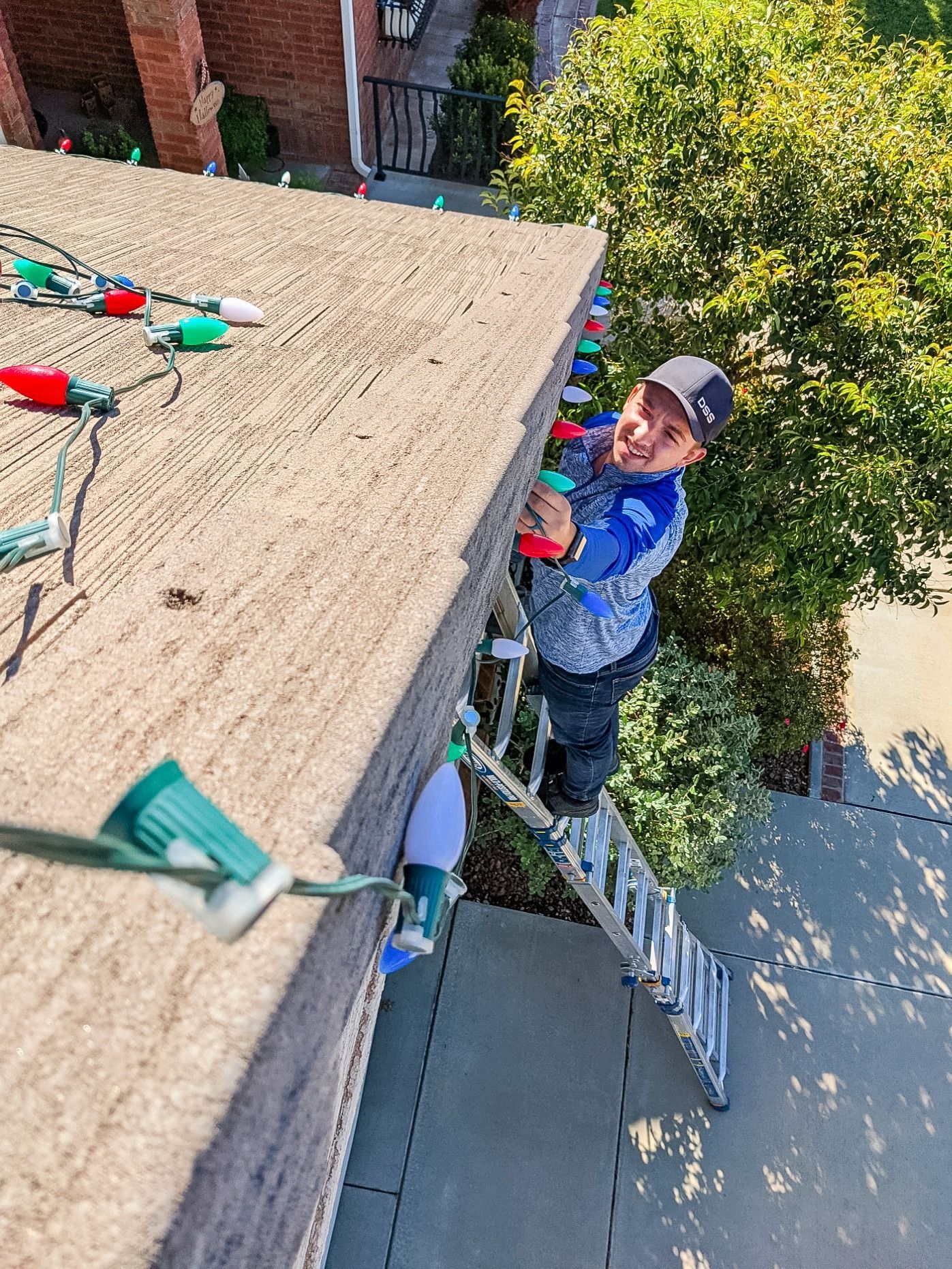 A person on a ladder hangs colorful holiday lights along the edge of a brick building's roof.
