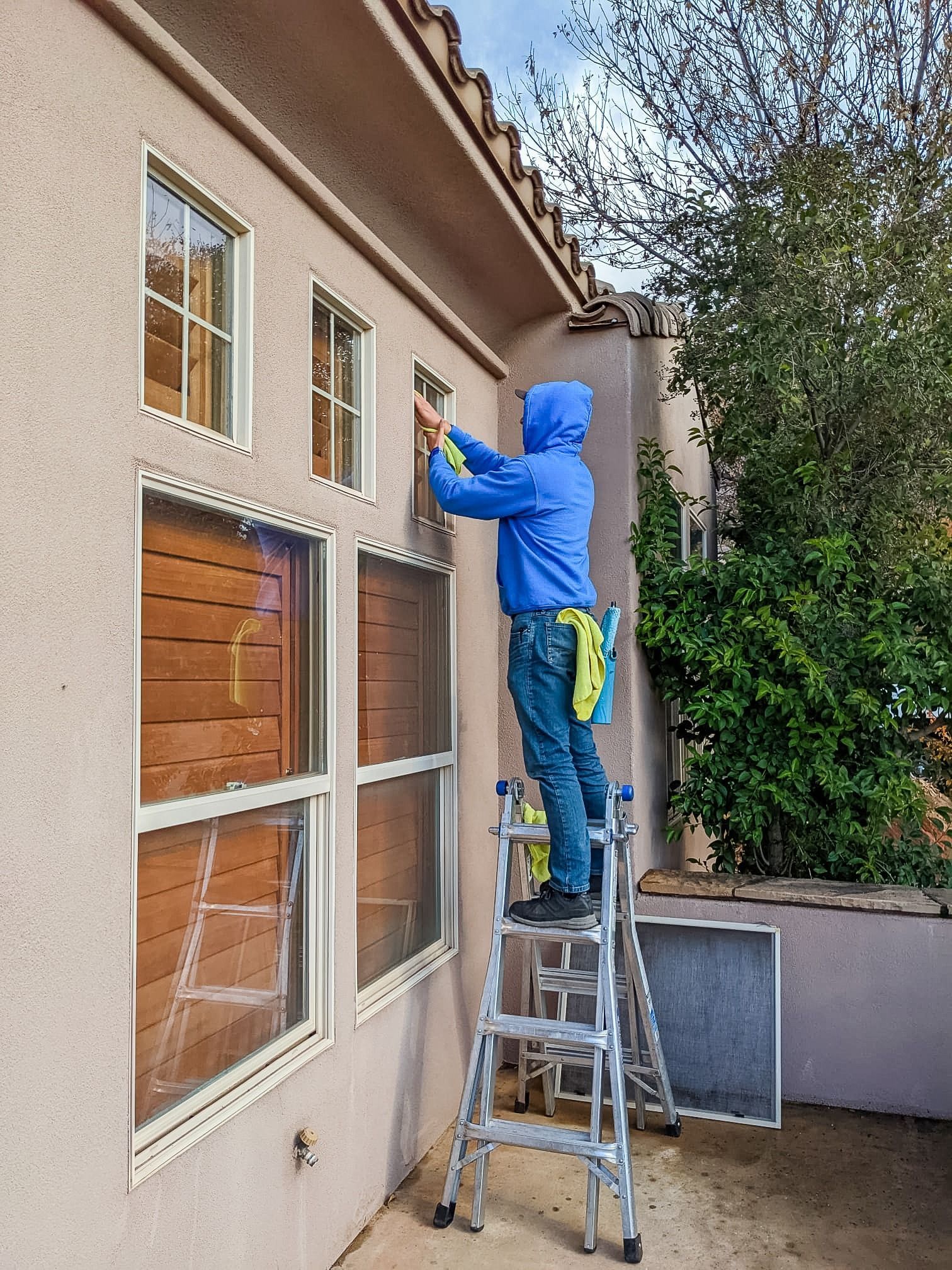 A person in a blue hoodie and jeans on a step ladder, cleaning the exterior of a high window on a stucco house.