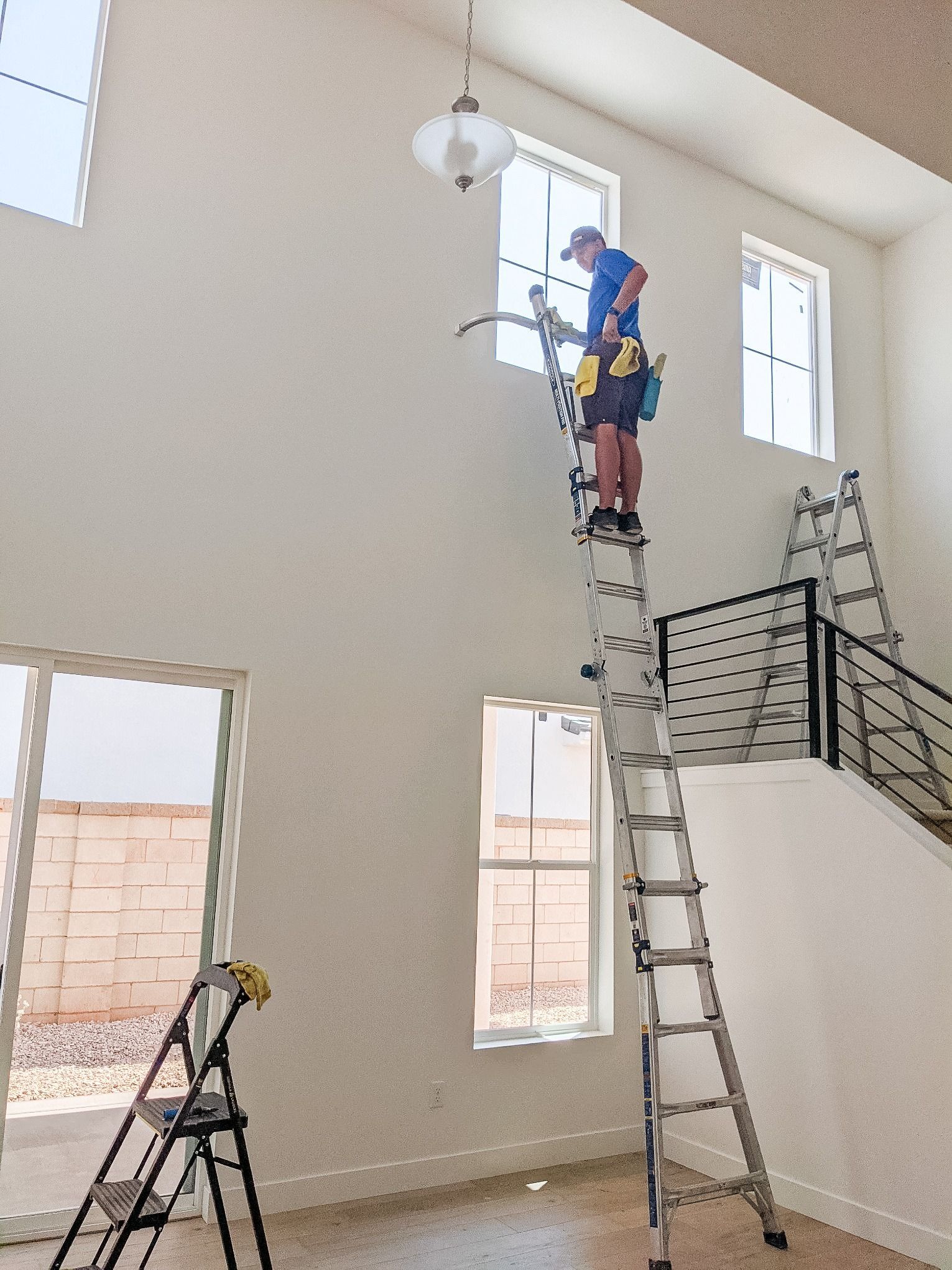 A person stands on a fully extended ladder to clean a high window in a modern home with a staircase nearby.