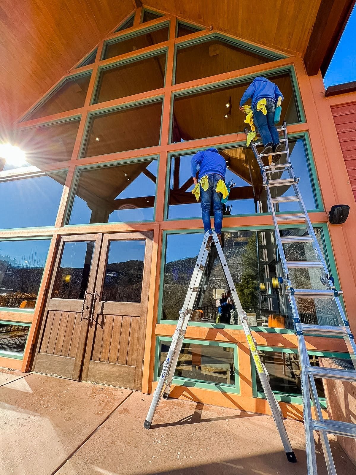 Two workers in blue uniforms clean the tall windows of a wooden building, using ladders on a sunny day.