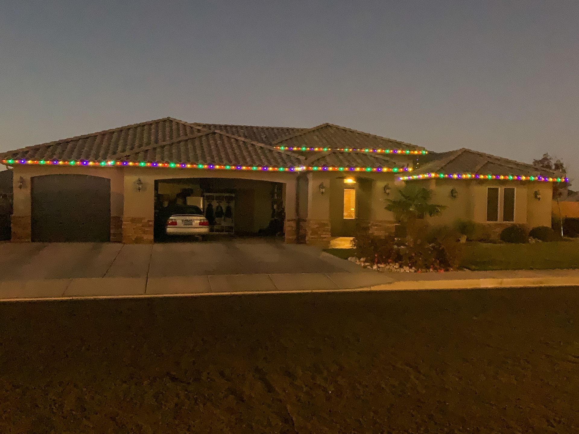 A single-story house at dusk with festive multi-colored holiday lights lining the roof eaves and a car parked in the garage.