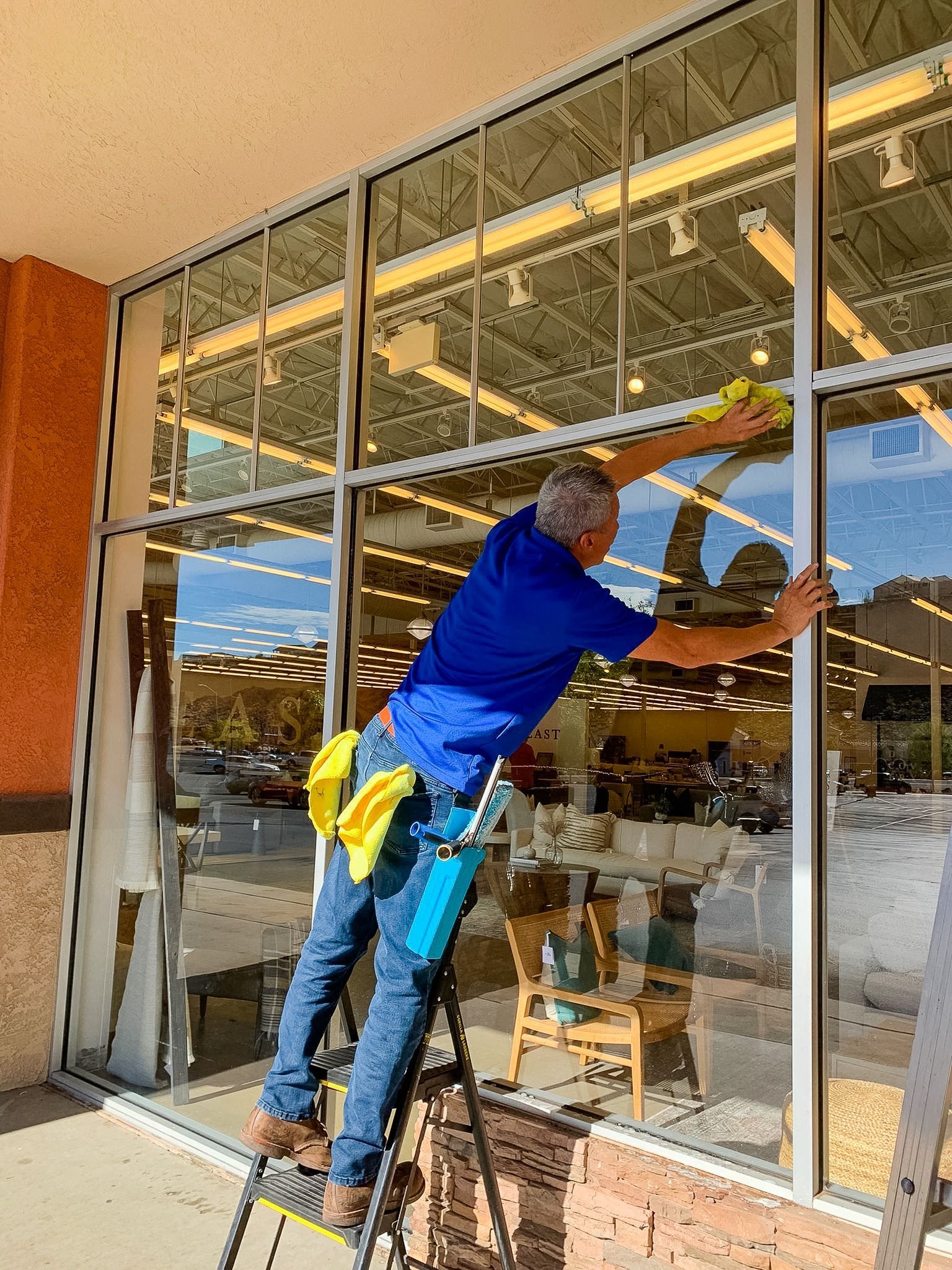 A person wearing a blue shirt and jeans stands on a ladder, using a yellow cloth to clean a large glass storefront window.