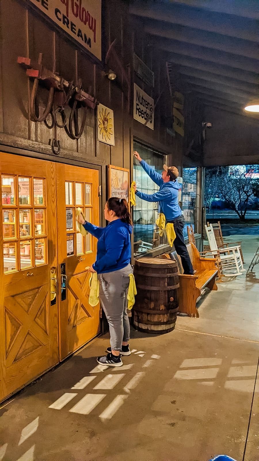 Two people wearing blue cleaning the windows of a rustic wooden storefront at dusk, with rocking chairs visible nearby.