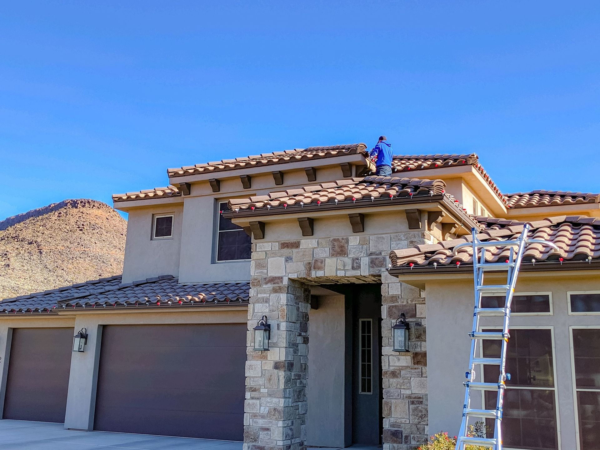 A person in a blue shirt works on the tiled roof of a tan, stone-accented suburban house, with a ladder leaned against it.