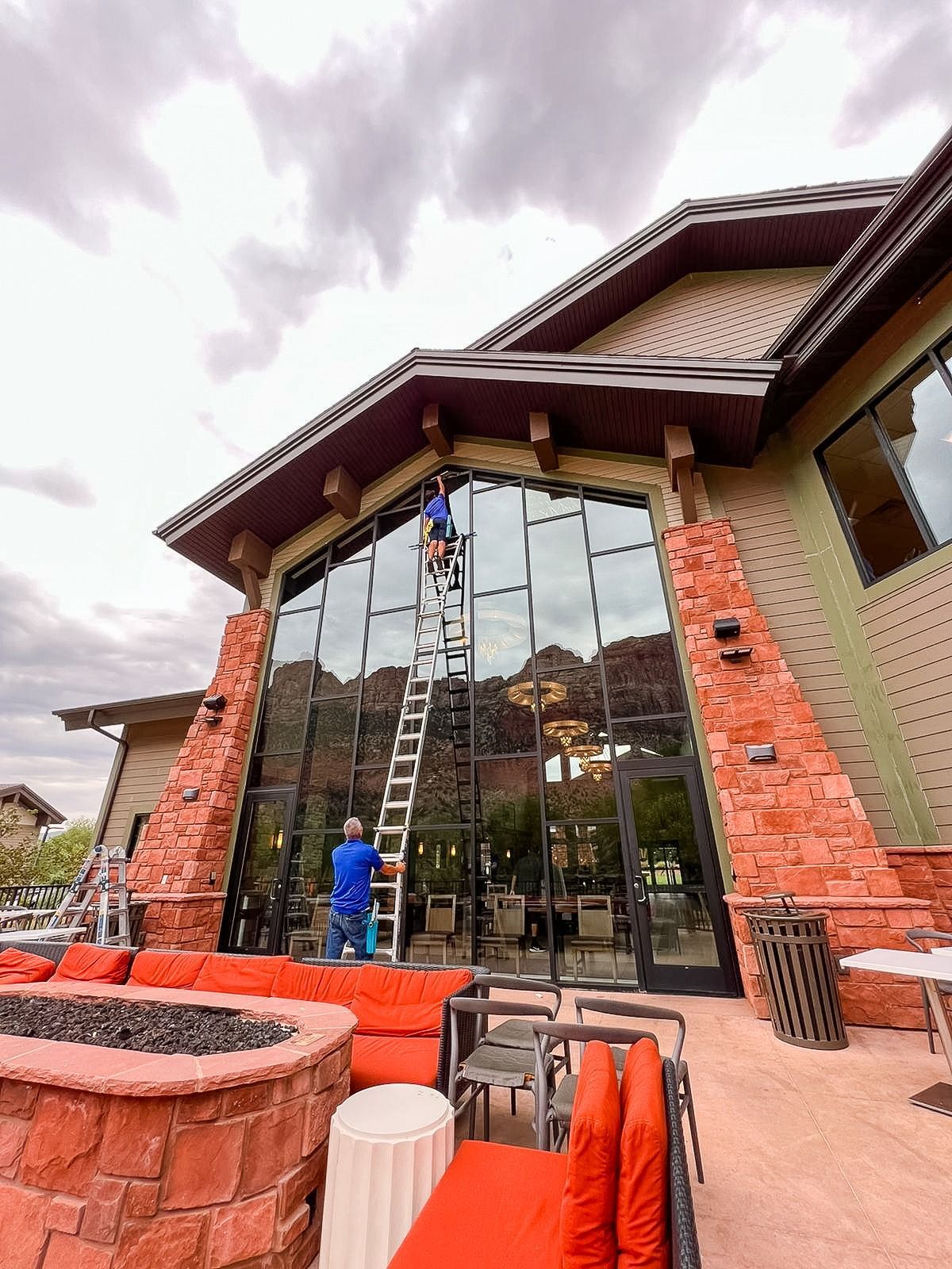 A worker on a tall ladder cleans the large, reflective glass windows of a building with red stone pillars and patio seating.