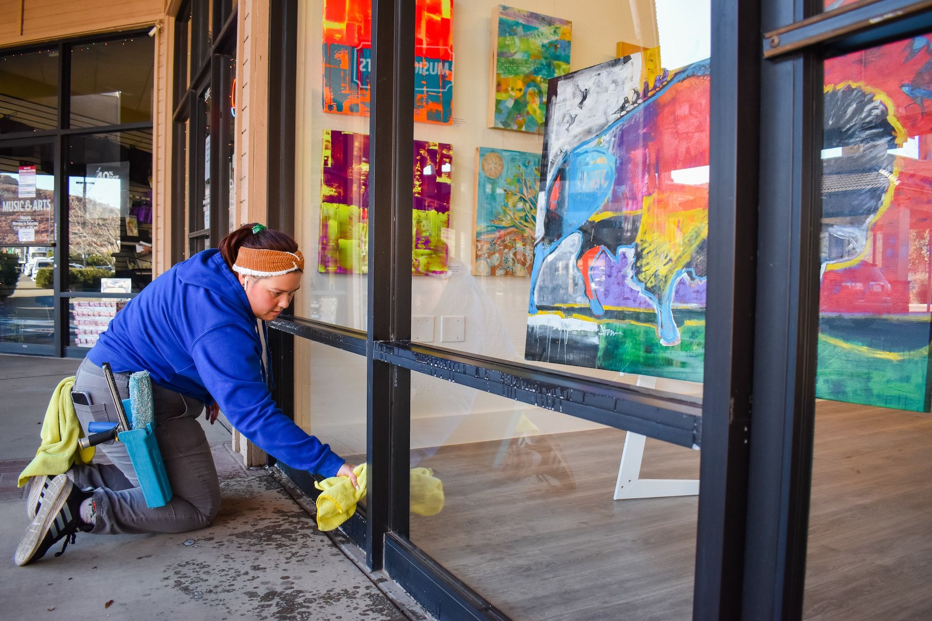 A person in a blue hoodie kneels while cleaning a large glass window outside an art gallery with colorful paintings.