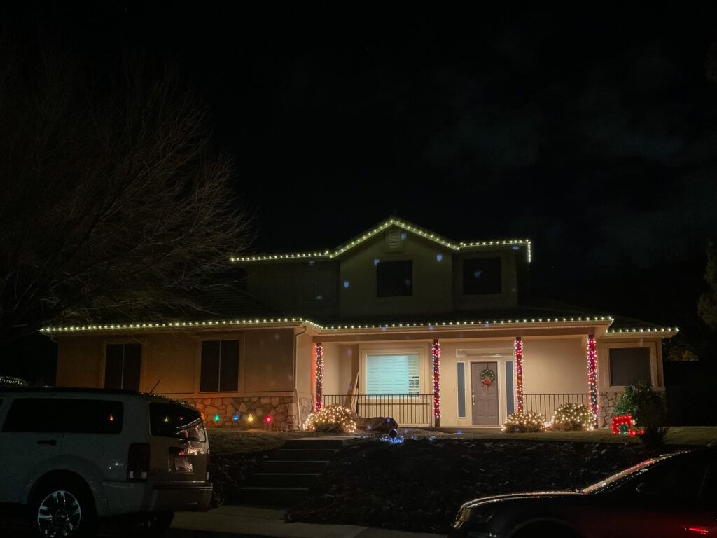 A house at night decorated with string lights along the roofline, pillars, and porch, with two cars parked out front.