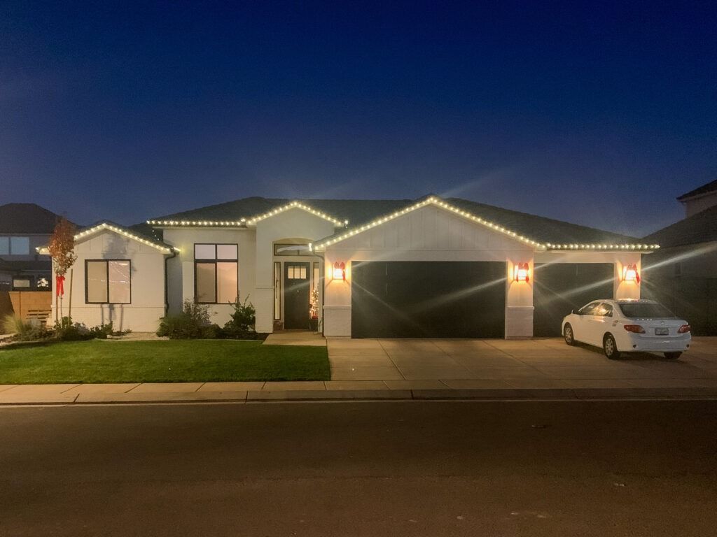 A single-story suburban home at twilight, decorated with white string lights along the roofline, with a car in the drive.