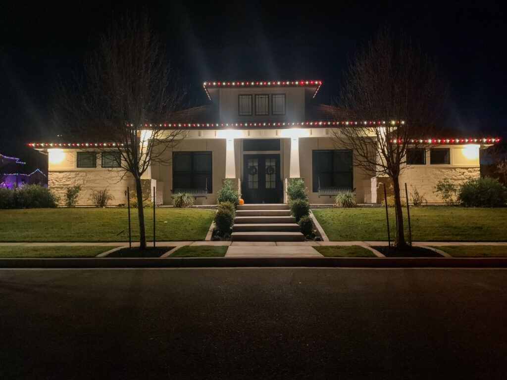 A single-story house at night illuminated by warm porch lighting and red roofline string lights, with a central staircase.