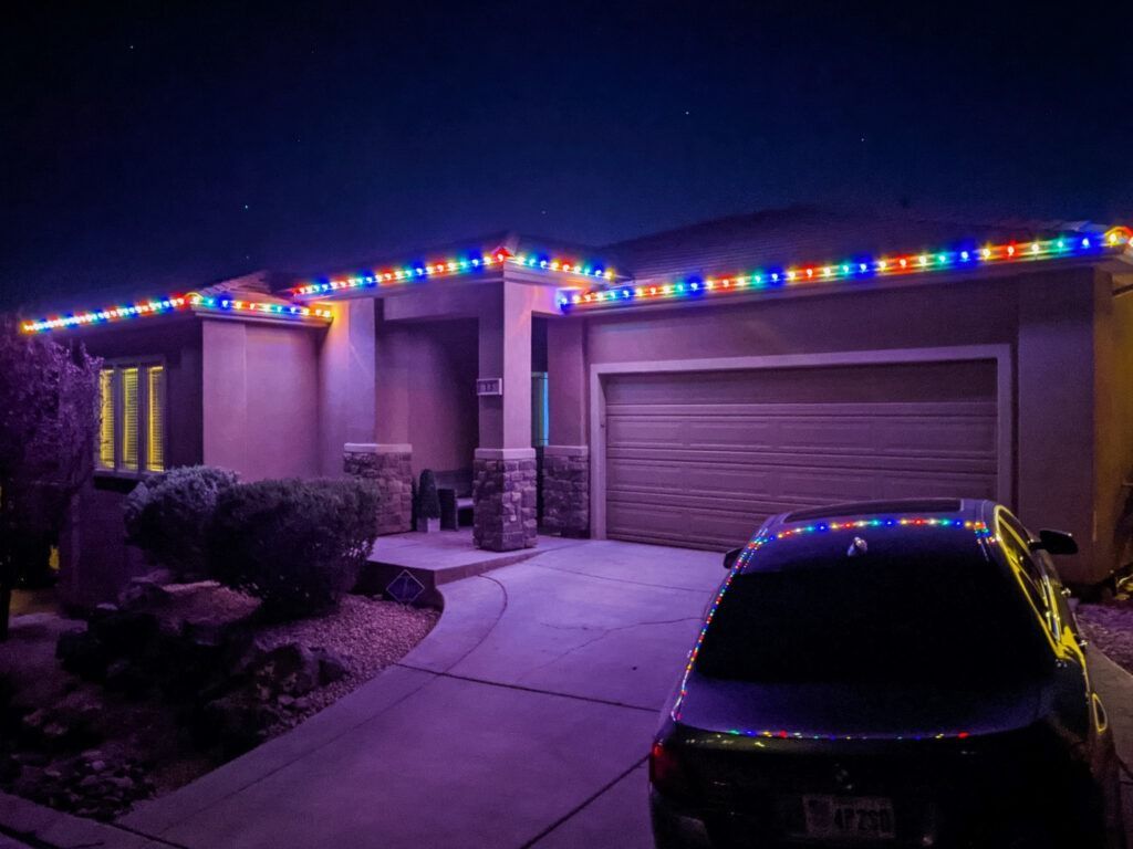 A suburban house at night illuminated with colorful LED holiday lights along the roofline, with a car in the driveway.