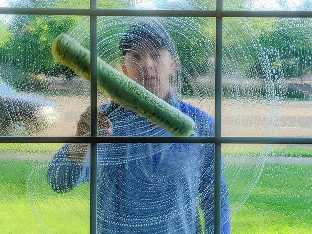 A person in a blue shirt cleans a pane of a divided window with a green-padded squeegee, creating circular soapy streaks.