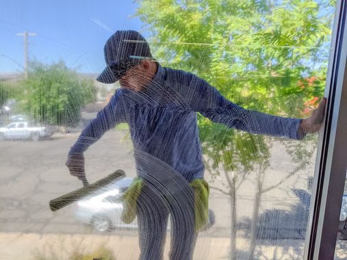 A person in a patterned shirt and hat uses a squeegee to clean a glass window overlooking a parking lot with trees.