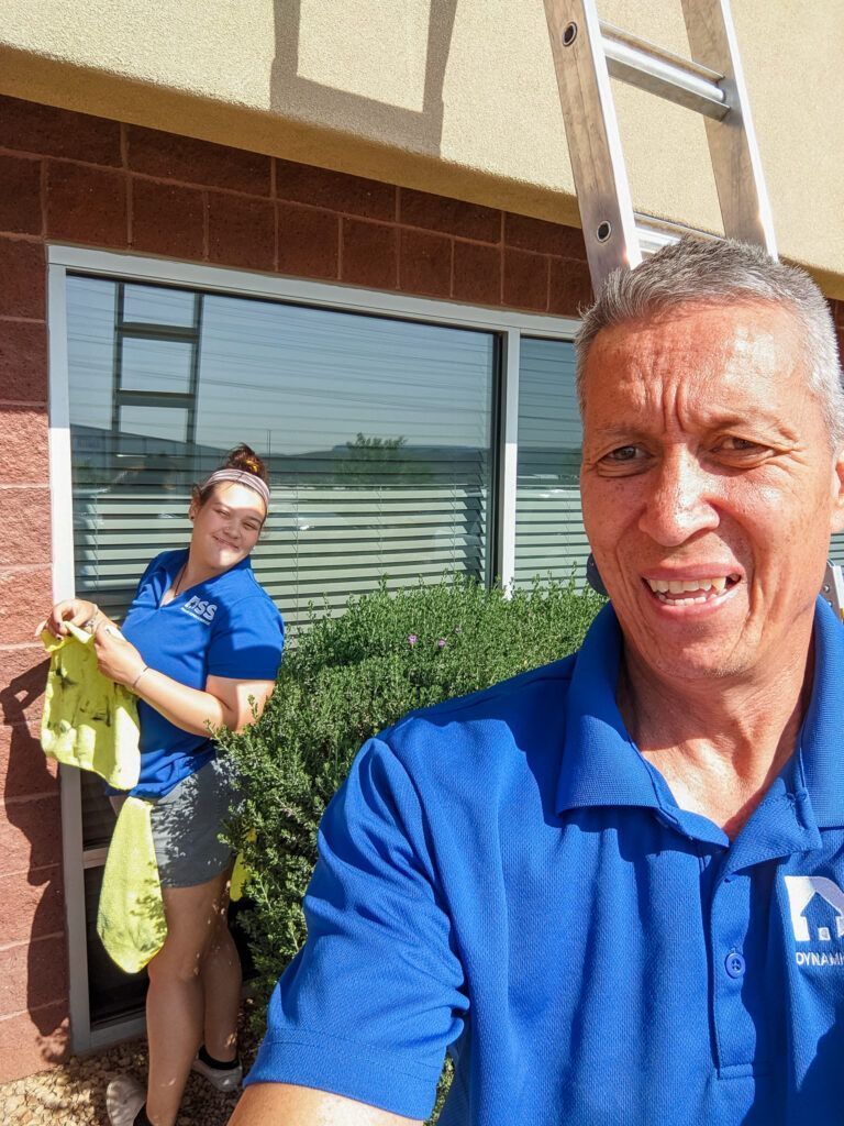 Two people in matching blue shirts and shorts stand outside by a building window, holding cleaning cloths near a ladder.
