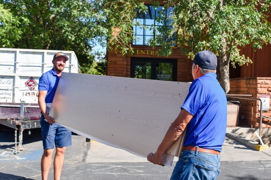 Two workers in blue shirts carry a large, rectangular piece of white construction material outside a building.