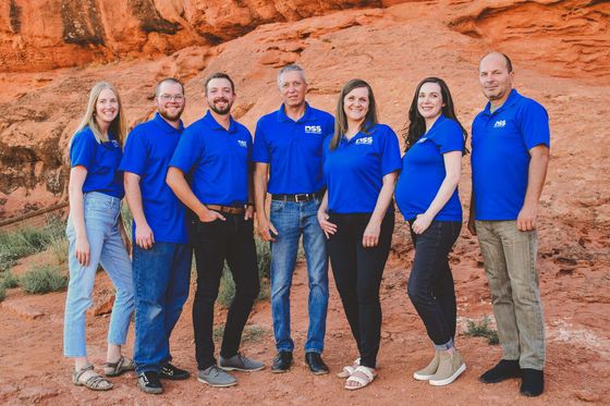 Seven people in matching blue polo shirts stand in a line against a reddish-brown rock formation.
