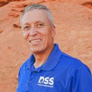 A smiling person in a blue polo shirt with a DSS logo, positioned in front of a red, rocky desert landscape.
