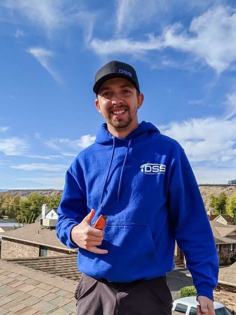 A smiling person in a blue hoodie and black hat gives a shaka sign while standing on a roof under a blue sky.