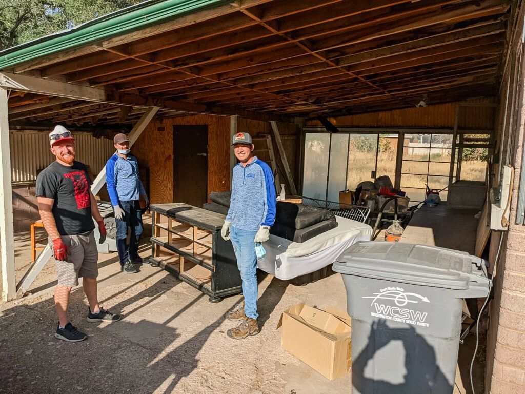 Three people stand inside an open-air shed filled with furniture and a gray recycling bin on a sunny day.