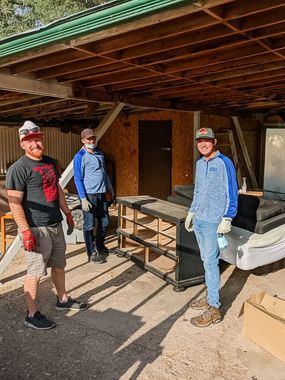 Three people wearing gloves and hats stand under a wooden carport structure, appearing to help with a cleanup or move.