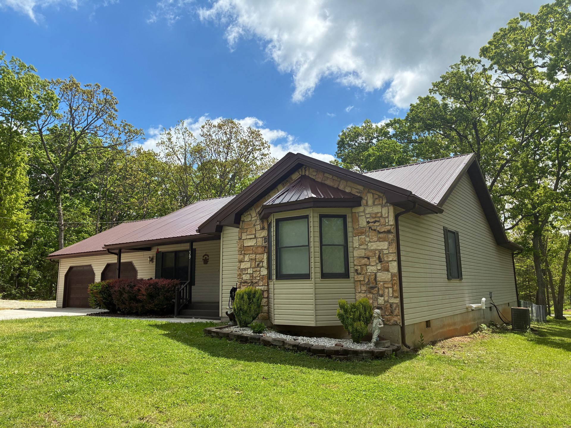 House with stone and tan siding, brown metal roof, and green lawn under a cloudy blue sky.