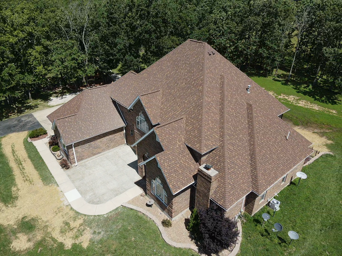 Aerial view of a brown-roofed house with brick accents, surrounded by greenery and a concrete patio.