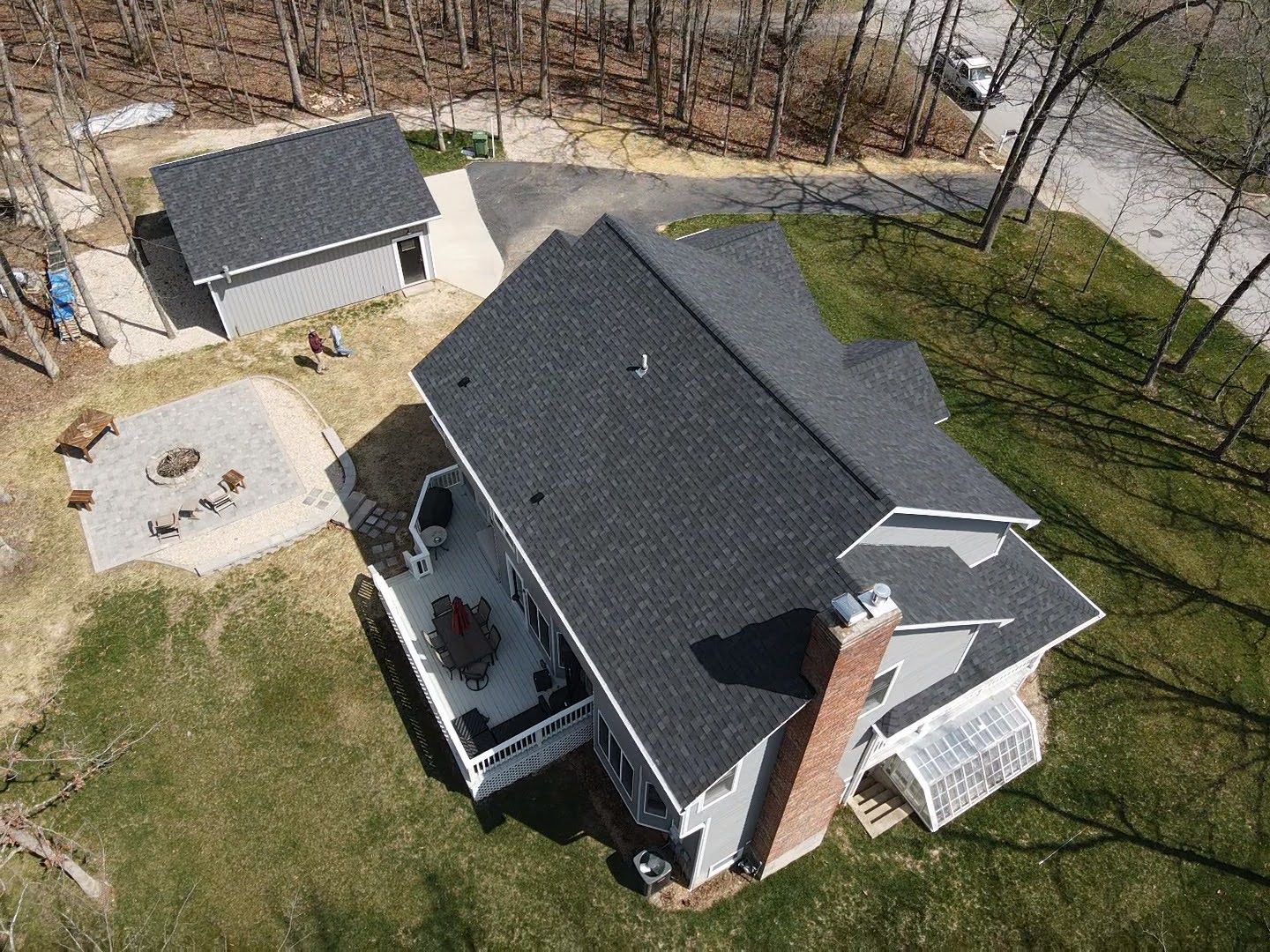Aerial view of a gray house with a dark gray roof, deck, chimney, and a detached gray garage in a wooded area.