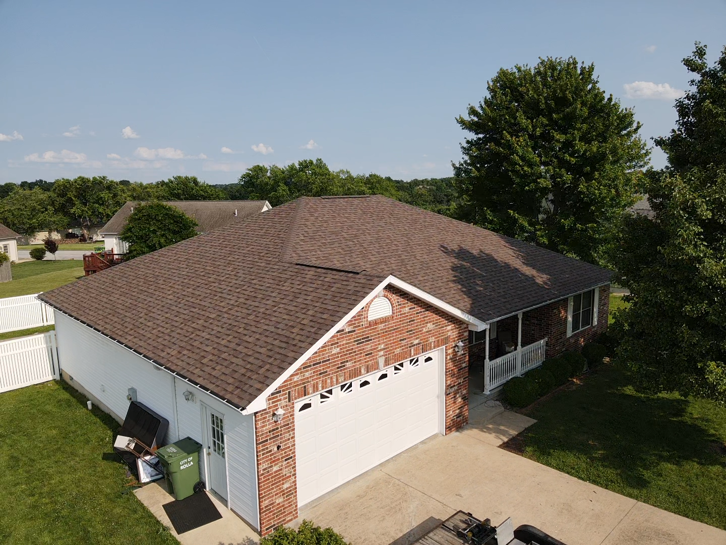 Brown-roofed house with brick facade and attached garage, on a sunny day. Green lawn and trees surround the structure.