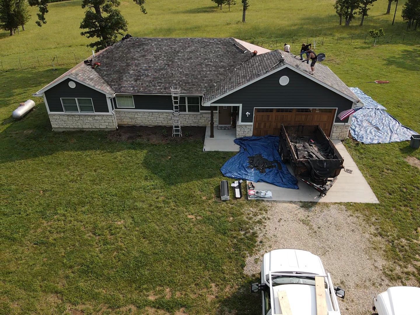 Roofers working on a house with a damaged roof, surrounded by tools, tarp, and a dumpster.