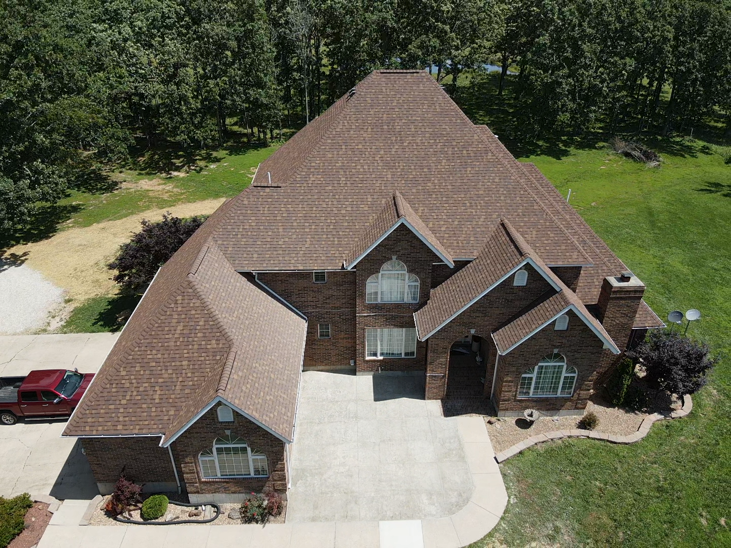 Brown-roofed brick house with a paved driveway, surrounded by green grass and trees.