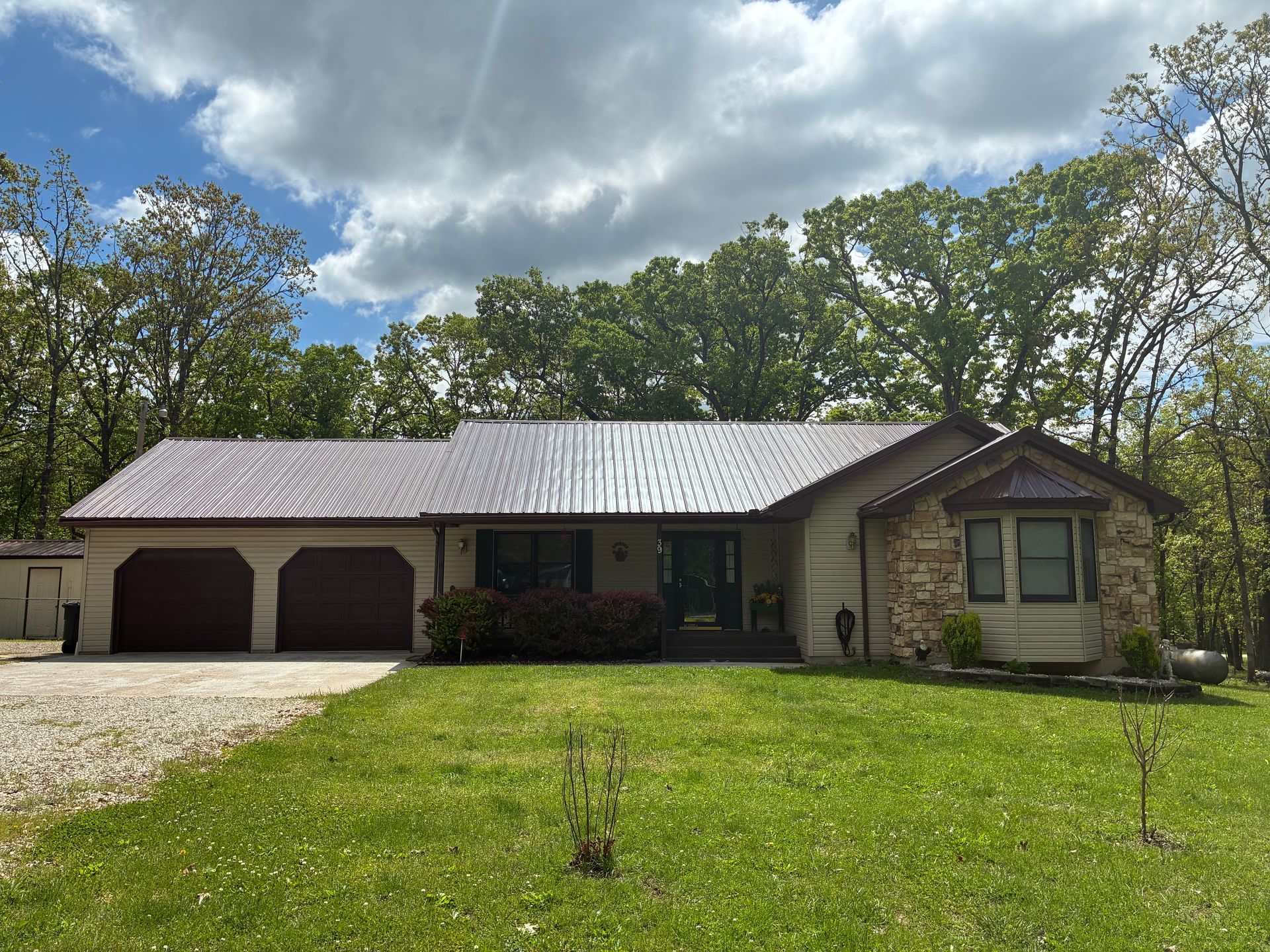 Tan house with brown roof, stone accents, and three-car garage in a wooded setting under a cloudy sky.
