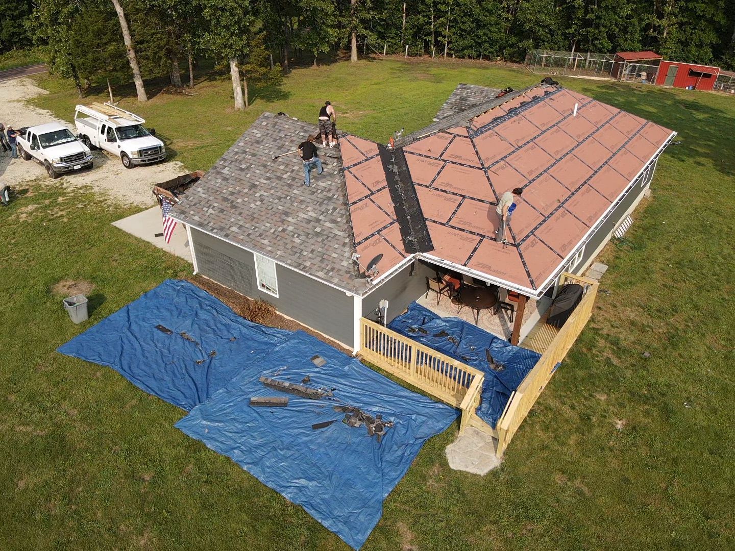 Roofers on a house with a partially shingled roof, some debris, two vehicles, and a blue tarp on the lawn.