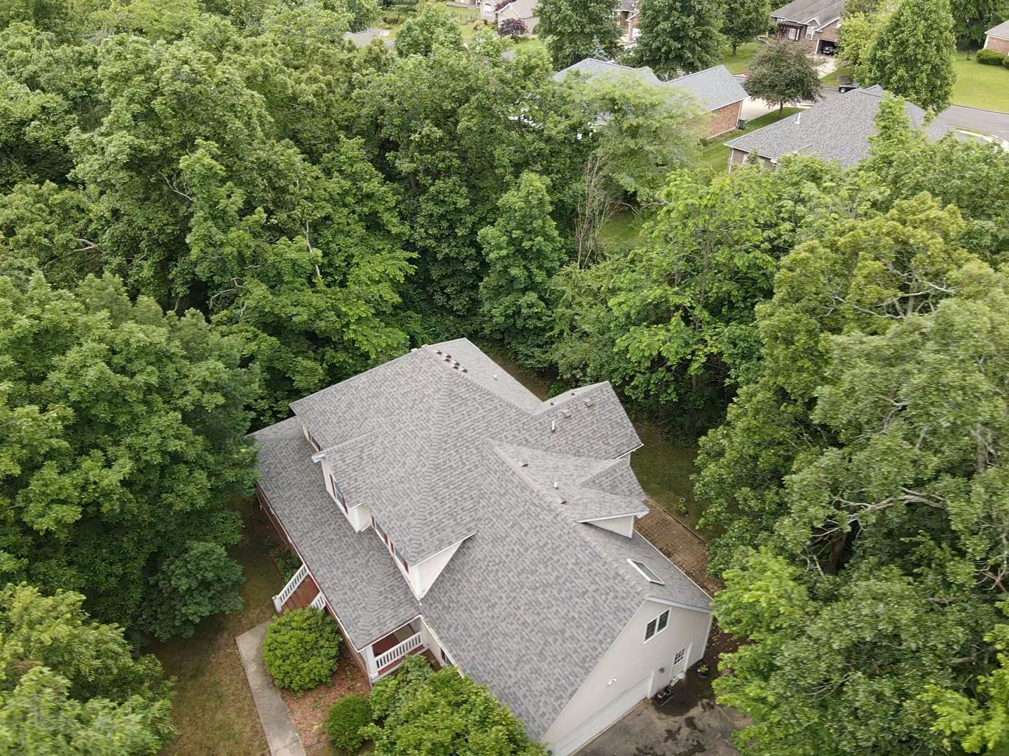 Overhead view of a house with a gray roof surrounded by green trees, with a few other houses visible in the background.