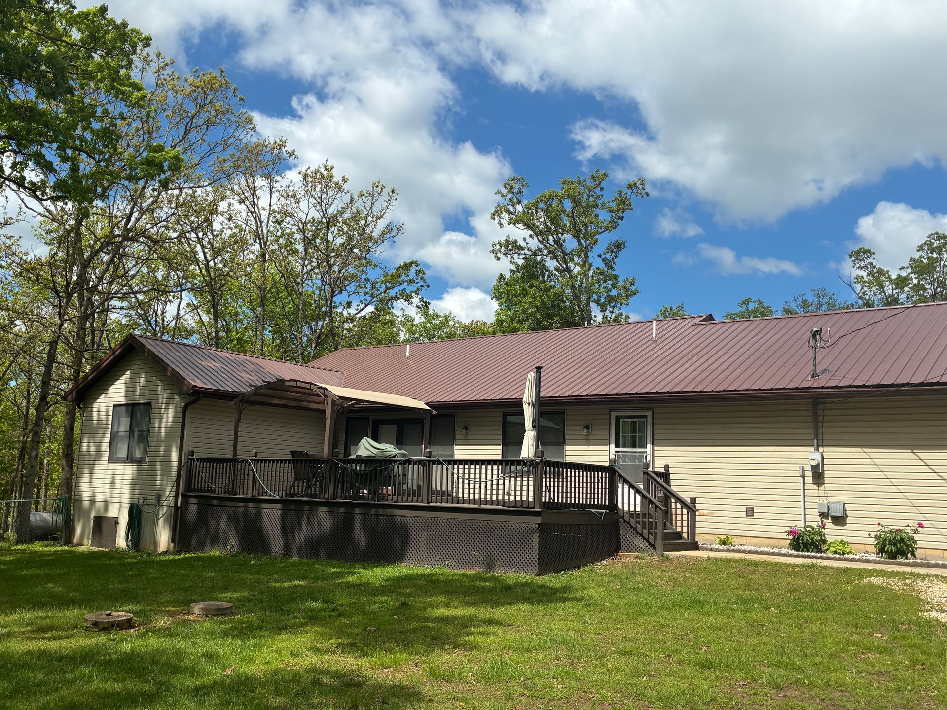 Brown-roofed house with deck, surrounded by trees, under a partly cloudy sky.