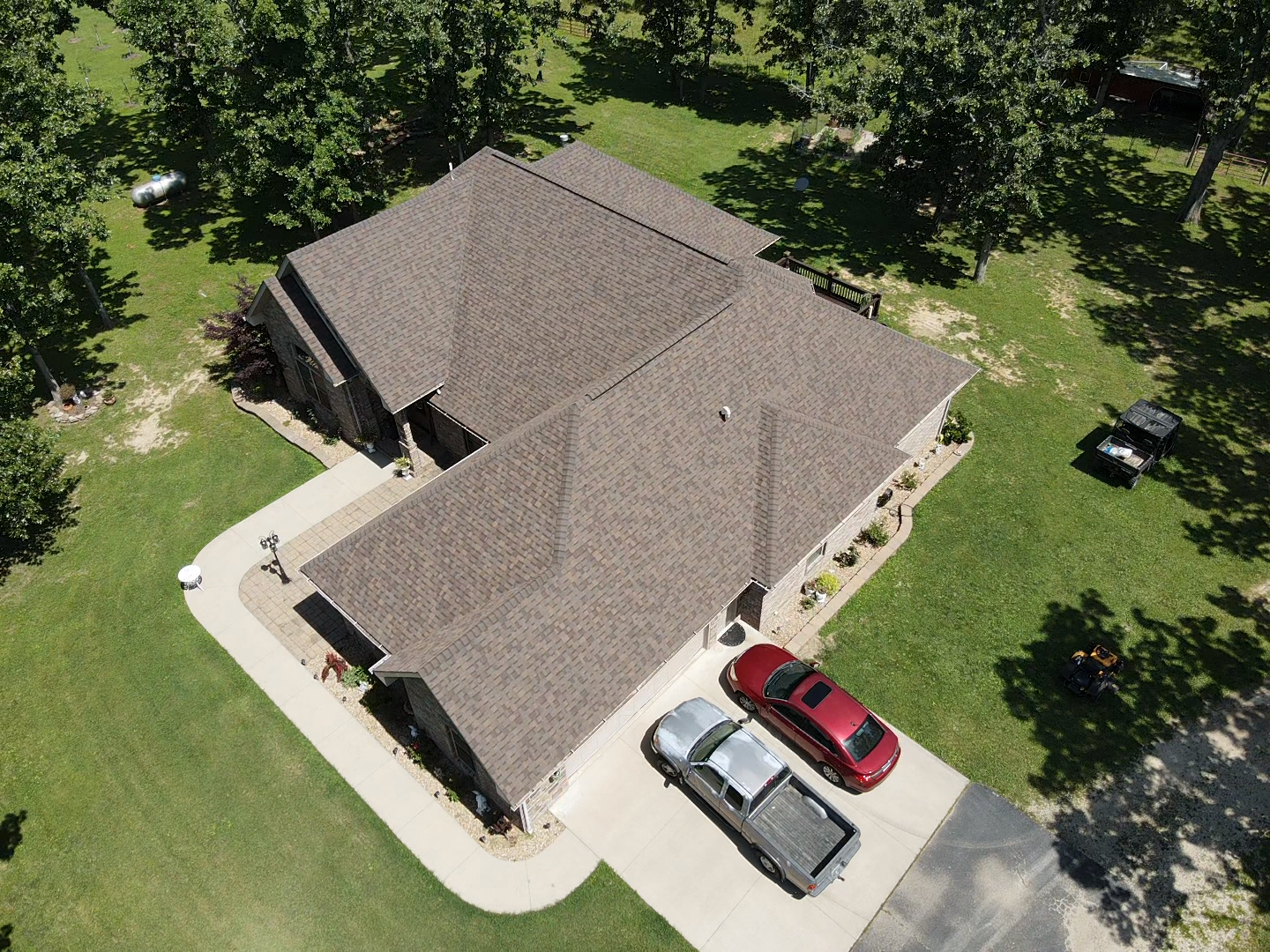 Aerial view of a brown-roofed house with two cars in the driveway, surrounded by green grass and trees.