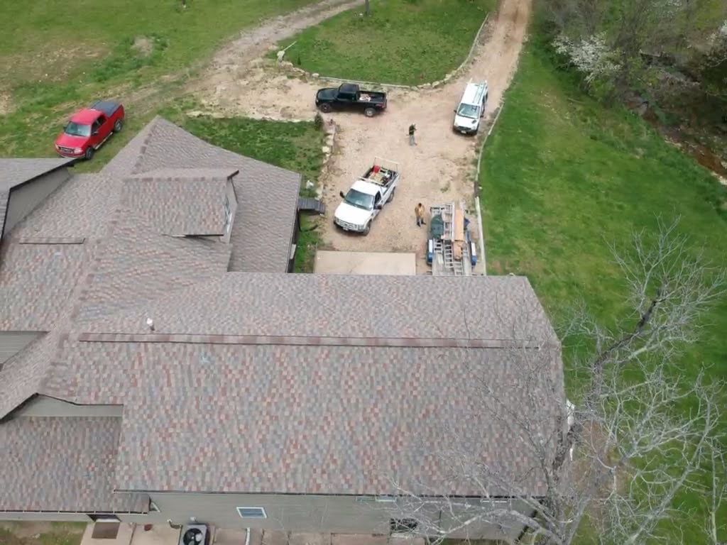 Aerial view of a house with a brown roof; several vehicles parked on a dirt road, person standing near.