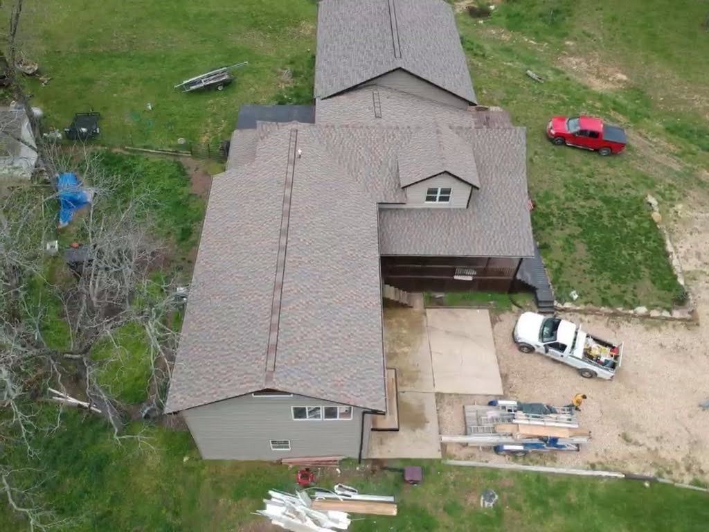 Aerial view of a house with a newly installed brown shingle roof, parked trucks, and construction materials.