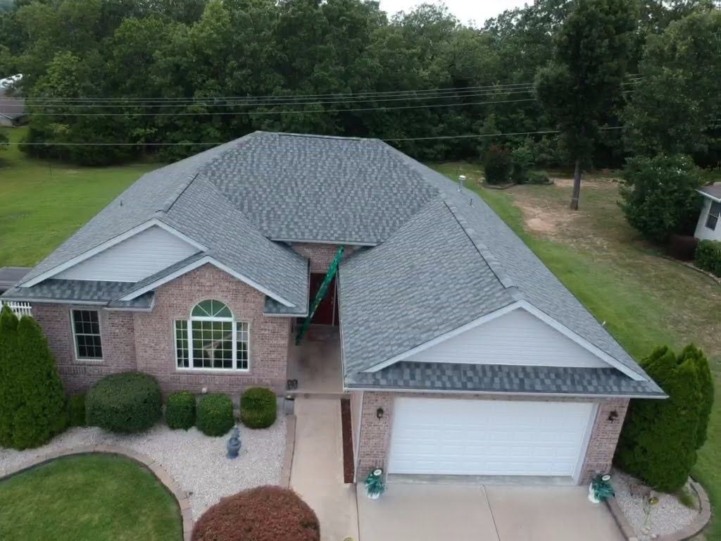 Aerial view of a brick house with a gray roof and a white garage door, surrounded by trees and landscaping.