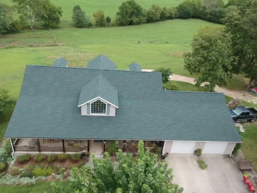 Overhead view of a house with a green roof, dormer, and attached garage, surrounded by a green field and trees.