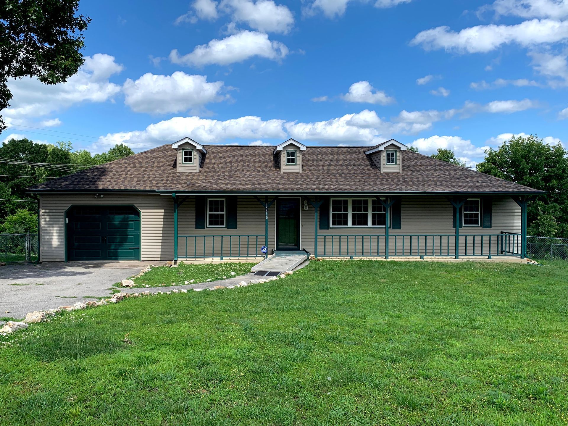 A tan one-story house with a green door and garage door sits on a grassy hill under a blue sky.